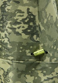 Zaļš kamuflāžas audums ar teksturētu virsmu. Satur Nike Tech birku, kas izstrādāta ar dažādiem rakstiem un zaļās krāsas toņiem.