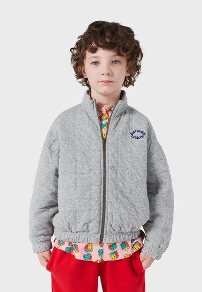 Bambino con capelli ricci che indossa una giacca grigia chiaro imbottita con zip, una maglietta a fantasia con dadi colorati e pantaloni rossi, in piedi con le mani in tasca.