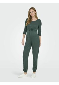 Combinaison verte à manches longues, taille élastique, jambes fuselées et en tissu doux. Portée avec des baskets blanches, se tenant devant un fond uni.
