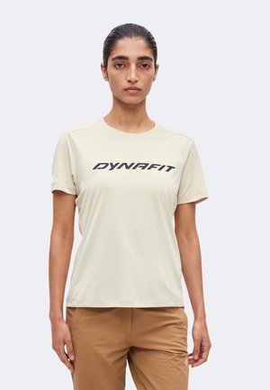 Γυναίκα με σκούρα μαλλιά μαζεμένα πίσω, που φοράει μια μπεζ μπλούζα Dynafit και ανοιχτό καφέ παντελόνι, στέκεται μπροστά σε απλό φωτεινό φόντο.