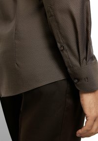 Braunes Hemd mit Muster und subtiler Diamantstruktur, mit geknöpftem Manschetten und geradem Saum, kombiniert mit dunkelbrauner Hose.