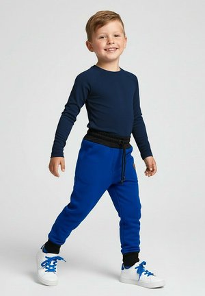 Uśmiechnięty młody chłopiec spacerujący, ubrany w granatową koszulkę z długim rękawem, niebieskie spodnie dresowe z czarnym paskiem i mankietami oraz białe tenisówki z niebieskimi sznurówkami.