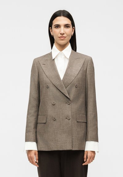 Femme aux longs cheveux foncés porte un blazer marron croisé sur une chemise blanche à col, debout devant un fond blanc uni.