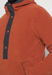 Pullover in pile color ruggine con cappuccio, caratterizzato da una patta bottonata blu navy e tasca frontale a marsupio; tessuto morbido e testurizzato.