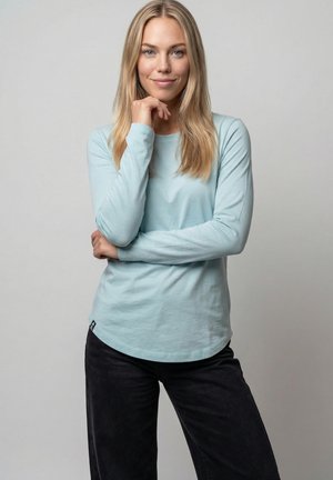 Frau mit langen blonden Haaren, die ein hellblaues Langarmshirt und eine schwarze Hose trägt, steht mit einer Hand nahe am Kinn vor einem einfarbigen Hintergrund.