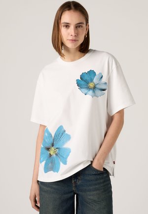 Femme portant un t-shirt blanc oversize avec deux grandes impressions de fleurs bleues, associé à un jean en denim foncé, debout devant un fond uni.
