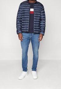 Granatowa kurtka puchowa z pikowanym wzorem, zestawiona z granatowym swetrem z czerwono-białymi paskami, jasnoniebieskimi dżinsami i białymi trampkami.