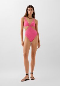 Roze gingham monokini met uitsnijding en cirkelaccent in het midden. Model gecombineerd met zwarte gestrikte sandaaltjes.