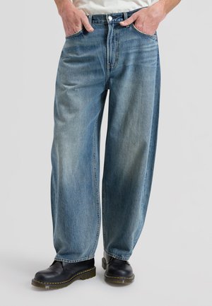 Henkilö, jolla on ylipitkät siniset farkut ja mustat nahkasaappaat keltaisella ompeleella, seisoo yksinkertaisen vaalean taustan edessä.