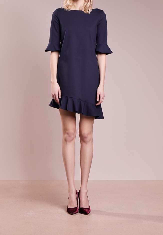 Robe bleu marine avec manches évasées et ourlet à volants. Tissu lisse et ajusté avec un décolleté carré. Portée avec des talons hauts bordeaux.