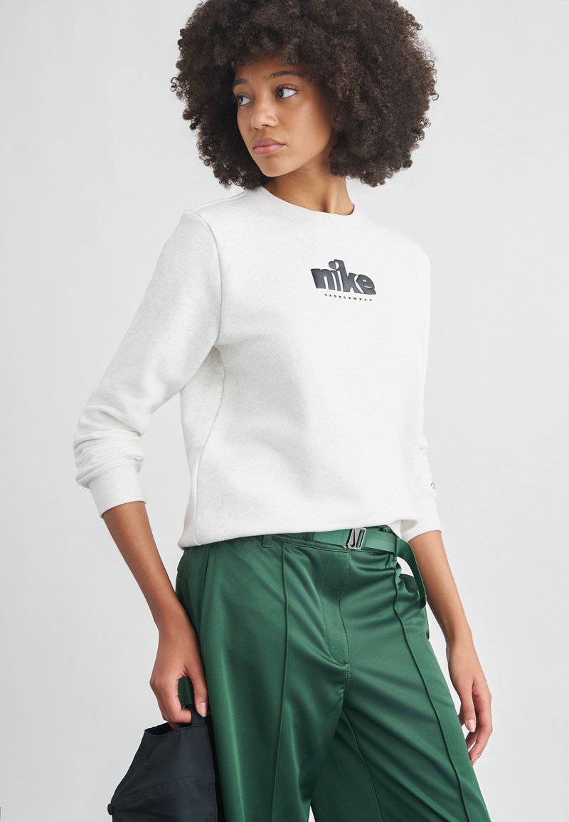 Νεαρή γυναίκα με σγουρά μαλλιά που φοράει λευκό φούτερ Nike και πράσινο παντελόνι, κρατώντας μαύρο καπέλο, κοιτάζοντας προς τα πλάγια.