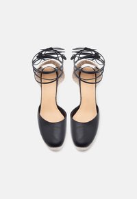 Ζευγάρι μαύρα δερμάτινα εσπαντρίγιες με μπεζ πλεκτές σόλες και μαύρα λουριά που δένουν στον αστράγαλο, εμφανίζονται από πάνω σε λευκό φόντο.