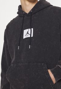 Felpa nera con texture sbiadita, tasca a kangaroo frontale, cordini regolabili e un'etichetta logo rettangolare bianca con una silhouette nera.