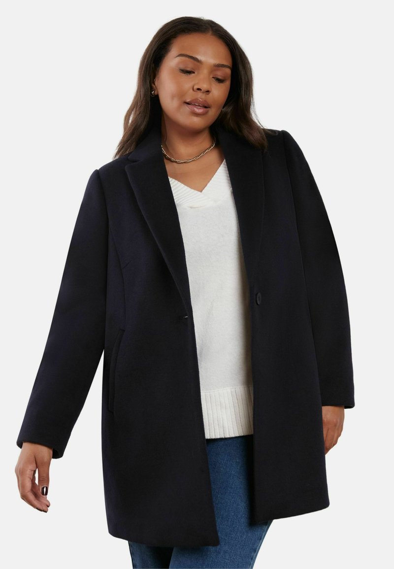 Marinblå ullrock med snedskuren krage, enkel knappstängning och sidofickor. Kombinerad med en vit tröja och blå jeans.