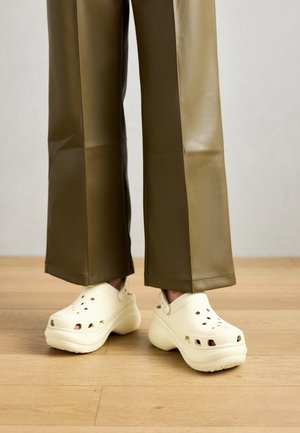 Personne portant des sabots compensés écru et un pantalon large vert olive, debout sur un sol en bois.