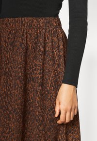 Taille en hand van een persoon die een zwarte geribde blouse met lange mouwen en een hoogoplopende bruine en zwarte patroonrok draagt.