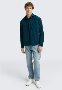 Tegenviolet corduroy overhemd met knoopsluiting, een enkele borstzak en lange mouwen, gecombineerd met lichtblauwe jeans en witte sneakers.