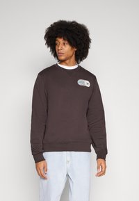 Dunkelbrauner Sweatshirt mit Rundhalsausschnitt, gerippten Bündchen und Saum, mit einem grafischen Patch auf der Brust. Kombiniert mit hellblauen Jeans.