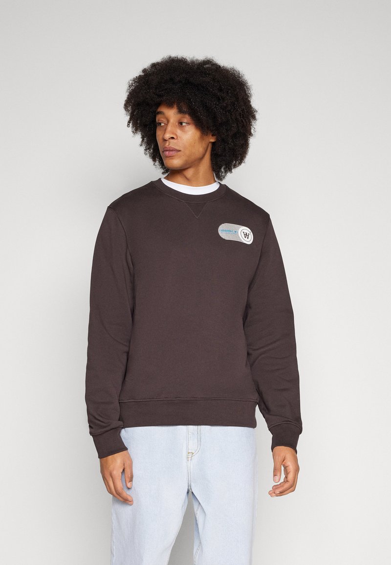 Dunkelbrauner Sweatshirt mit Rundhalsausschnitt, gerippten Bündchen und Saum, mit einem grafischen Patch auf der Brust. Kombiniert mit hellblauen Jeans.