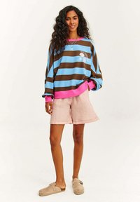 Pruhovaný modro-hnědý oversized svetr s růžovými manžetami, doplněný o světle růžové lněné šortky a béžové slip-on boty.