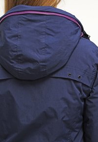 Giacca blu navy con cappuccio, con zip a contrasto viola e dettagli con occhielli. Tessuto liscio e leggero con finitura opaca.