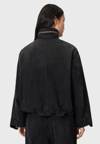 Veste en denim noir avec un col haut, des épaules tombantes, un ourlet élastique et une texture lisse. Présente un détail de couture contrastant.