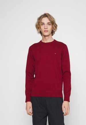 Maglione rosso a maglia con scollo rotondo, maniche lunghe e un piccolo logo sul petto. Il materiale sembra morbido e strutturato.