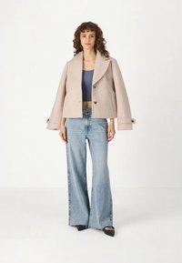 Μπεζ κοντό μπουφάν με ζακάρ καπίτ (notched lapels), μπροστινά κουμπιά και πλάγιες τσέπες, συνδυασμένο με ανοιχτό μπλε φαρδιά τζιν παντελόνια. Μαύρες επίπεδες γόβες.