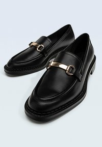 Mocassins en cuir noir avec une finition lisse, présentant un accent en métal doré et des détails en couture sur une semelle contrastée.