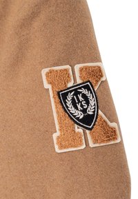 Tessuto beige con una patch con la lettera "K" in rilievo marrone e beige, sovrapposta da uno scudo nero con una corona di alloro bianca e il testo "IKKS".