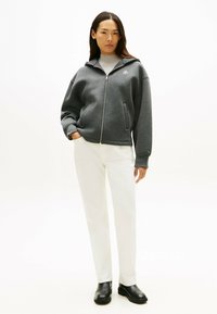 Un sweat à capuche gris zippé avec une coupe décontractée et des poches latérales, associé à un pantalon blanc et des bottines noires, présenté sur un fond neutre.