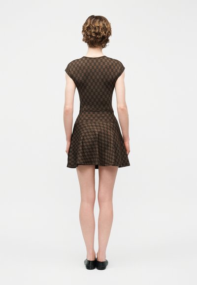 Γυναίκα με κοντά σγουρά μαλλιά που φοράει εφαρμοστό, καφέ φόρεμα κοντών μανικιών με σχέδιο και μαύρα παπούτσια, όρθια με την πλάτη γυρισμένη σε απλό φόντο.