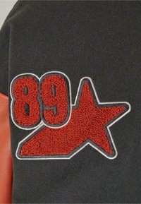 Chiffres « 89 » en chenille rouge et écusson étoile bordé de blanc sur tissu gris foncé, partie d’un vêtement avec détail en cuir marron.