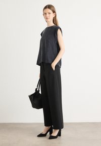 Top senza maniche grigio scuro, pantaloni larghi neri, tacchi neri, che tiene una borsa a secchiello nera. Tessuto morbido e leggero con un taglio informale.