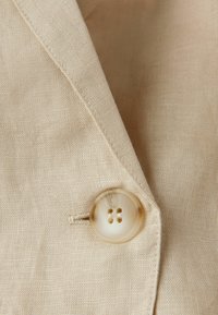 Béžová lněná tkanina s detailním záběrem na kulatý čtyřdírový knoflík s hladkým povrchem, umístěný na texturovaném pozadí.