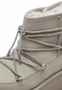 Bottine beige avec cuir texturé, doublure en fausse fourrure et lacets décoratifs en argent. Présente des bords courbés et une semelle épaisse.