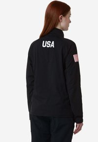 Giacca nera con colletto alto e maniche lunghe. Presenta la scritta bianca "USA" e una patch con la bandiera americana sulla manica sinistra. Texture liscia.