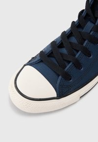 Nærbilde av en marineblå kanvas sneaker med svarte snører og en hvit gummitåhette på en lys bakgrunn.