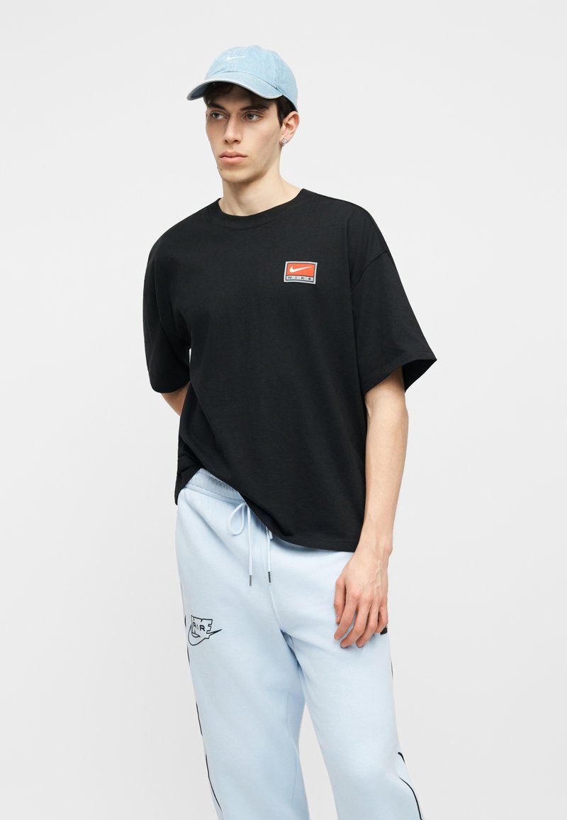 Giovane uomo con indosso una t-shirt nera Nike, pantaloni della tuta Nike azzurri chiari con coulisse e un cappellino azzurro chiaro, in piedi contro uno sfondo neutro.