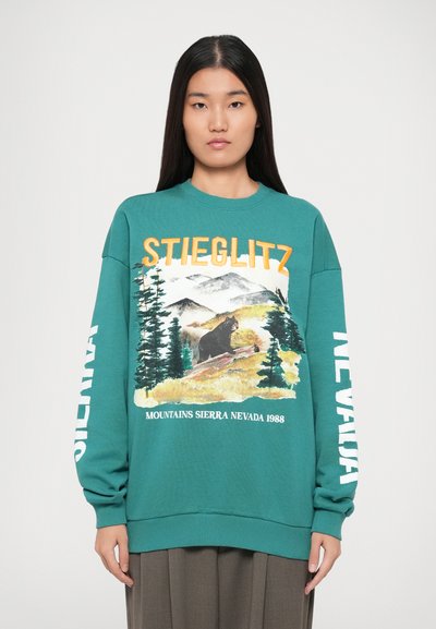 Sweatshirt vert d'eau avec un imprimé de paysage montagneux et une illustration d'ours. Présente un texte blanc : "STIEGLITZ" et "MOUNTAINS SIERRA NEVADA 1988."