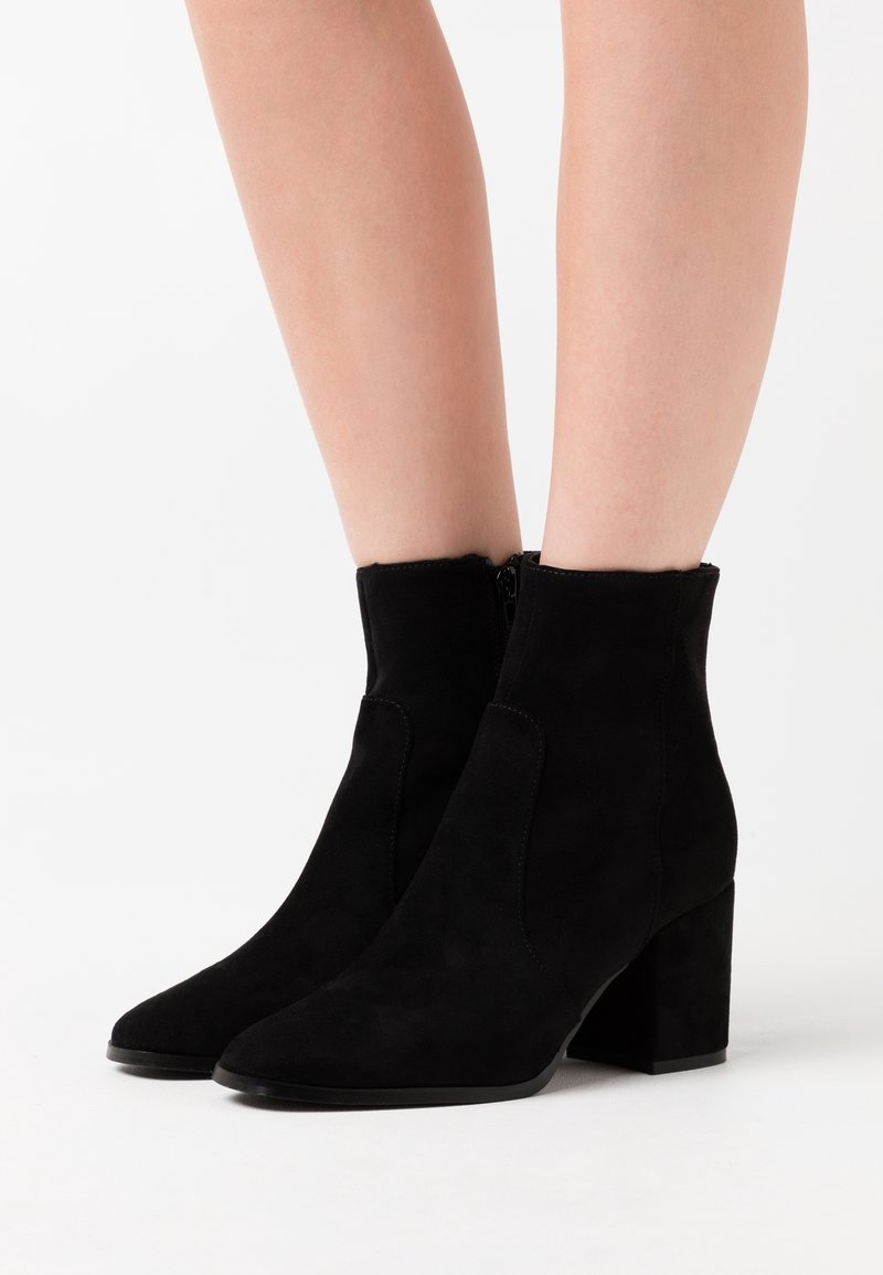 Jambes portant des bottines en daim noir avec talons blocs et fermetures éclair latérales sur fond blanc.