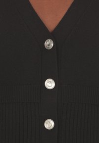 Cardigan nero con scollo a V, texture a coste e bottoni argentati con dettaglio del logo. Il materiale sembra morbido e aderente.