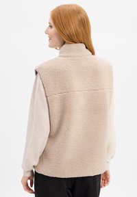 Beżowa kamizelka z polaru z wysokim kołnierzem, bez rękawów, o teksturowanej powierzchni, z miękką podszewką, noszona na jasnym, długim rękawie.