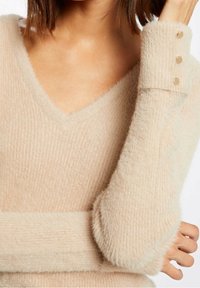 Pull beige col en V avec texture côtelée et matière douce et duveteuse. Doté de boutons décoratifs aux poignets pour plus de détail.