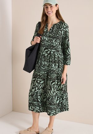 Donna sorridente che indossa un vestito midi verde e nero con motivo a fantasia, cappellino verde chiaro, scarpe beige e tiene una borsa tote nera sulla spalla, su sfondo neutro.