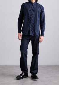 Marineblaue Hemd mit Bandkragen, kombiniert mit dunklen Jeans und glanzschwarzen Schuhen, die einen geraden Schnitt und dezente Textur aufweisen.