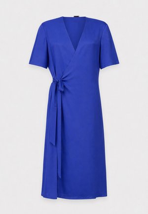 Woman wearing a blue wrap dress with short sleeves, black sunglasses, and silver pointed mules against a plain white background.