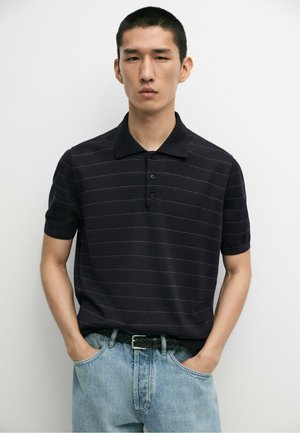 Jeune homme aux cheveux noirs courts portant un polo rayé foncé et un jean bleu clair, debout les mains dans les poches.