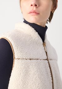 Cream-colored shearling vest with a beige trim, featuring a zip closure and a high collar. Worn over a navy turtleneck.