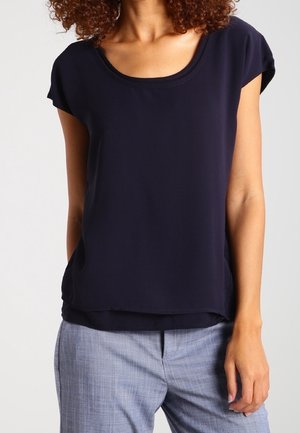 Kvinde iført en mørkeblå kortærmet bluse med rund halsudskæring og lysegrå skræddersyede bukser mod en ensfarvet baggrund.